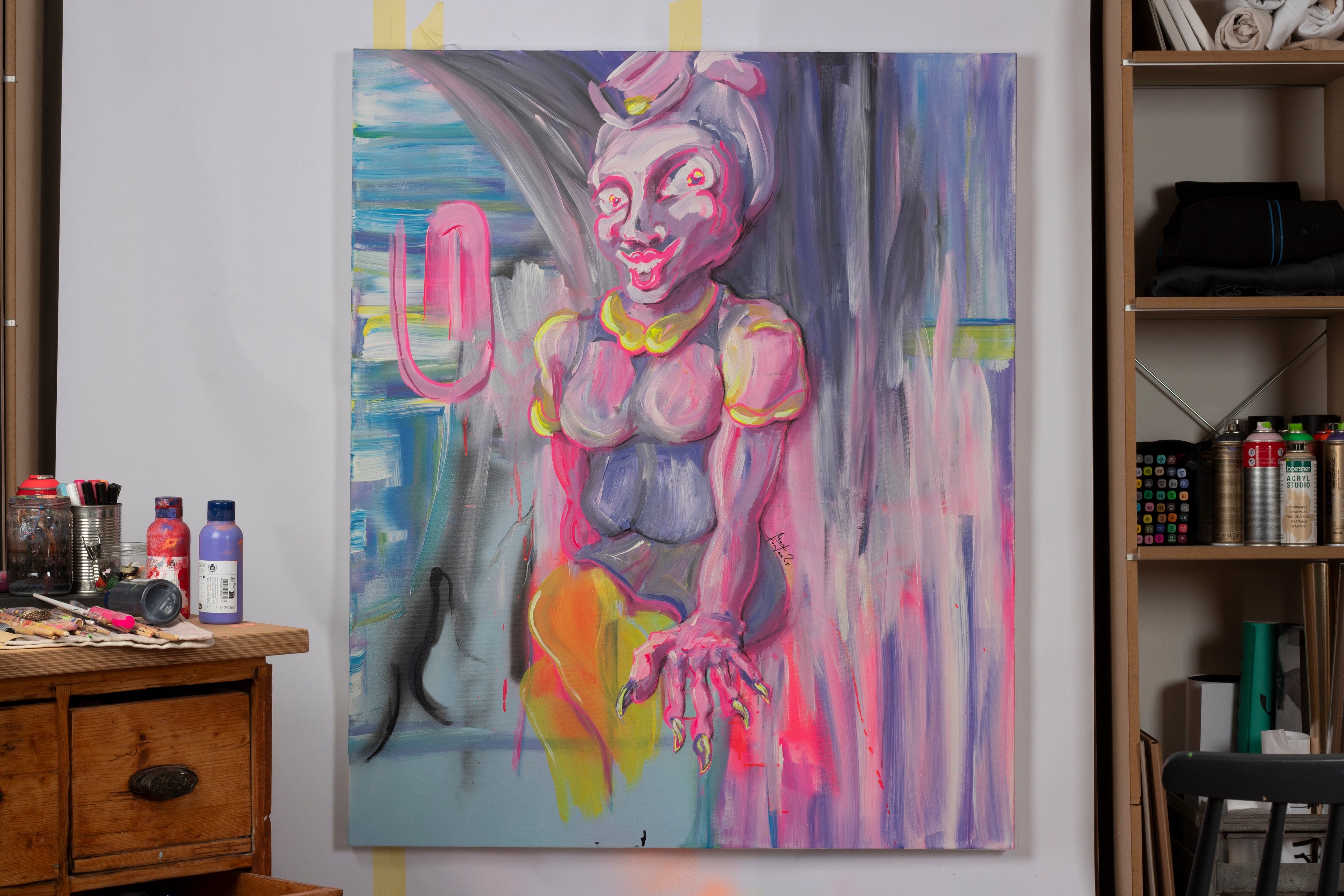Zeitgenössisches figuratives Gemälde einer stilisierten weiblichen Figur mit überzeichneten Gesichtszügen und Neonakzenten – expressive Malerei zu Identität, Inszenierung. Sitzende Figur in Rosa-, Grau- und Gelbtönen mit betonten Augen und Händen vor abstrakt verlaufendem Hintergrund mit gestischen Pinselspuren in Blau, Grau und Pink im Atelier mit Spraydosen, Farbtuben, Pinseln und Markern präsentiert.