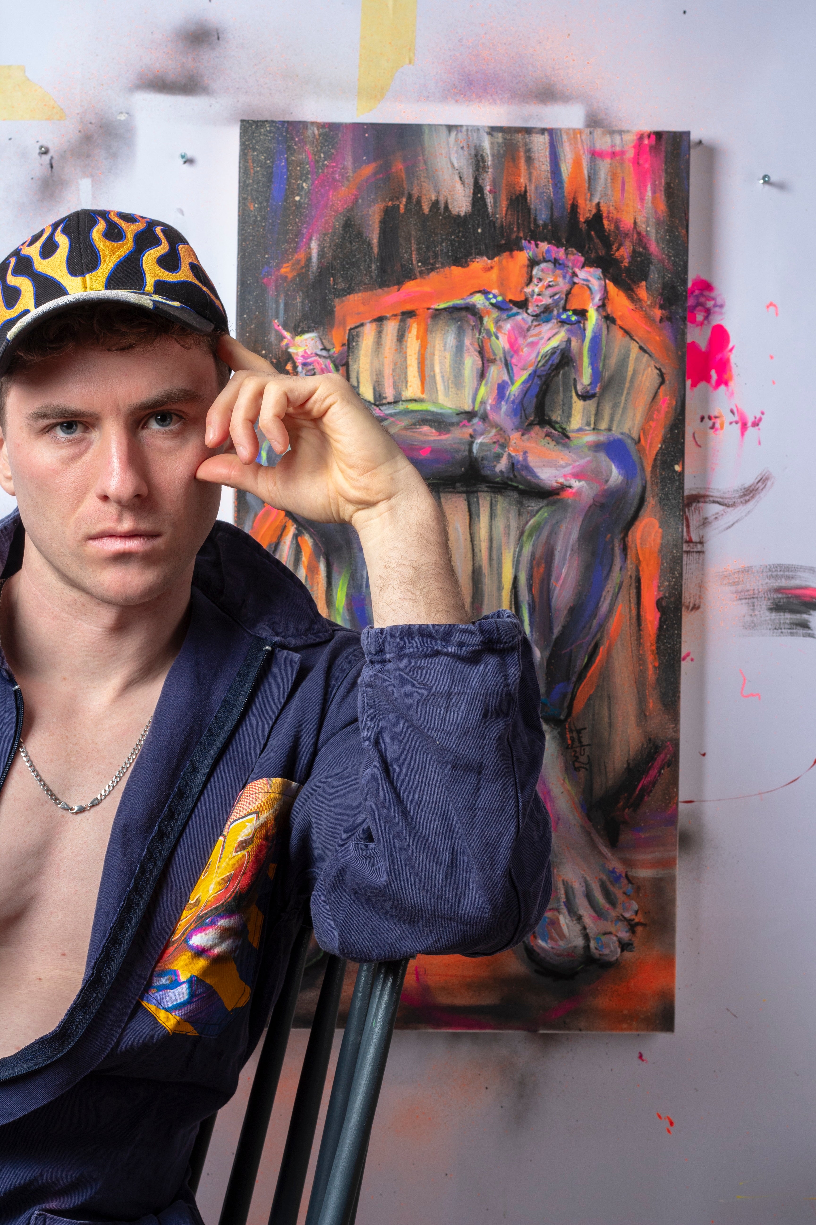 Zeitgenössisches Künstlerporträt im Atelier mit expressivem figurativen Gemälde (TRE KRONOR) im Hintergrund – visuelle Inszenierung zu Identität, Autorenschaft und künstlerischer Praxis. Nah dargestellte Person mit Cap mit Flammendekor und direktem Blick vor farbintensiver Malerei einer sitzenden Figur mit Neonakzenten in Orange, Pink und Violett auf roh bearbeiteter Atelierwand.